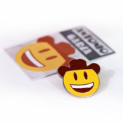 Photo of a enamel pin with yellow, brown and white smiley face with a cowboy hat.  silver sides can be seen.  blurred packaging can be seen in the background on a white background.