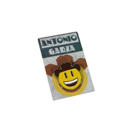 Photo of a enamel pin with yellow, brown and white smiley face with a cowboy hat, on a white background..  silver sides can be seen.  enamel pin is on top of packaging that says "Antonio Garza" on it. 